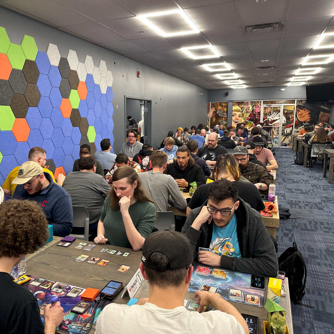 A large indoor event space filled with people seated at long tables playing card games. The room has gray walls, a colorful hexagonal wall paneling on the left, and bright overhead fluorescent lighting.