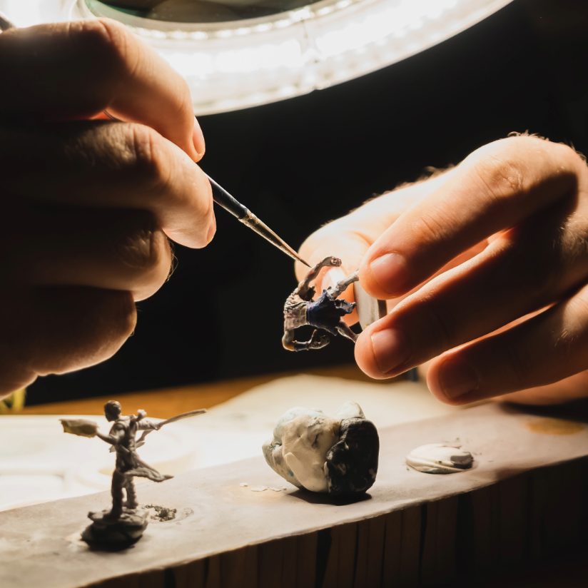 Close-up of a person's hands using tweezers to work on a small miniature figurine on a workbench under a ring light.