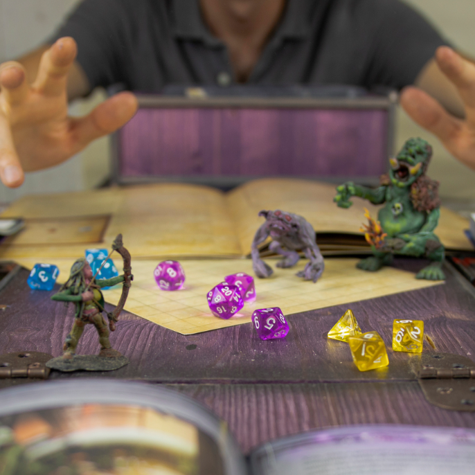 A tabletop gaming scene with a person's hands and upper torso visible behind a desk, with open books, dice, and small fantasy figurines in the foreground. The product appears to be tabletop gaming accessories or miniatures shown in use.